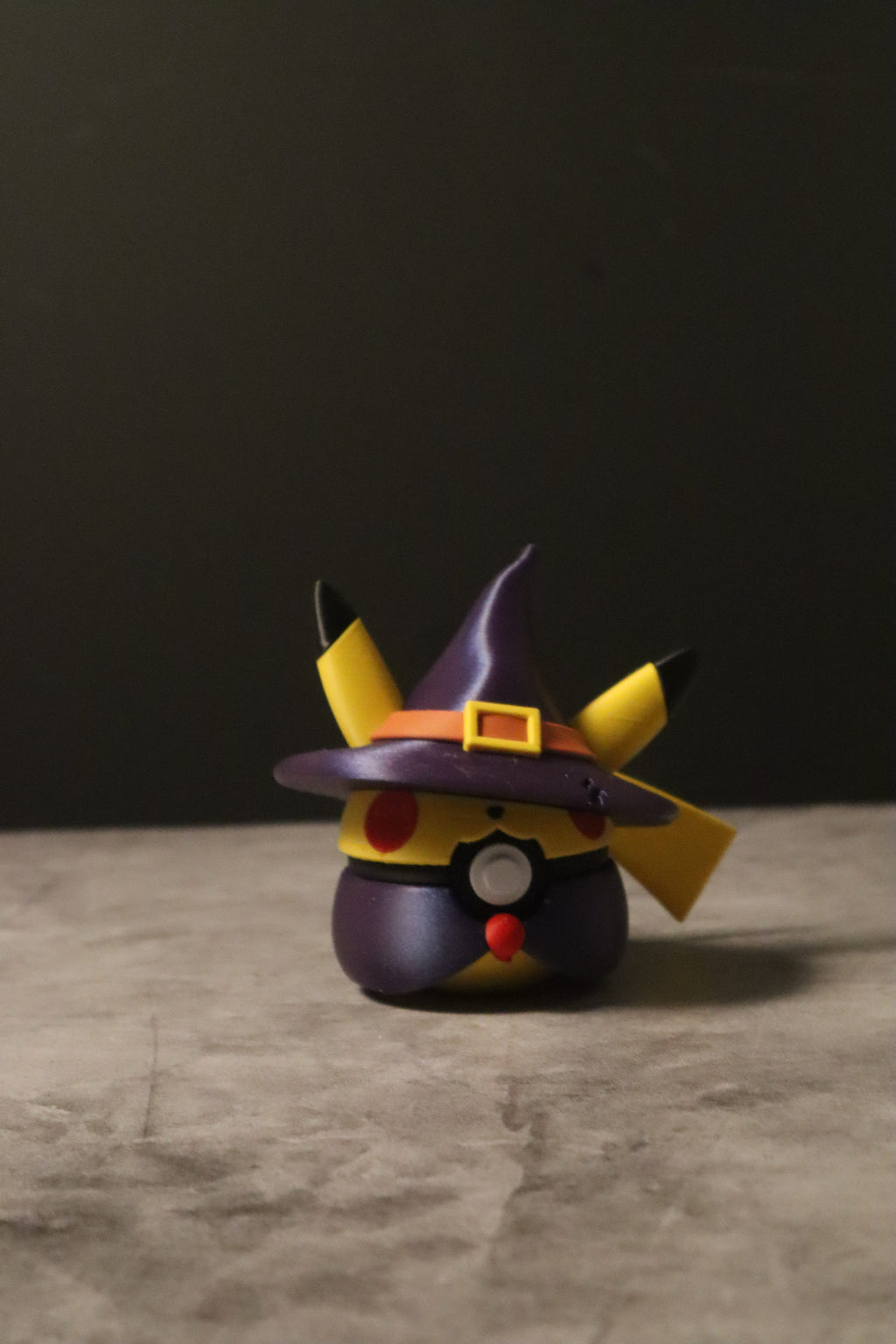 A small Pikachu-themed character figurine wearing a purple witch hat with yellow ear tips, standing on a textured gray surface against a dark background.