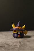 A small Pikachu-themed character figurine wearing a purple witch hat with yellow ear tips, standing on a textured gray surface against a dark background.