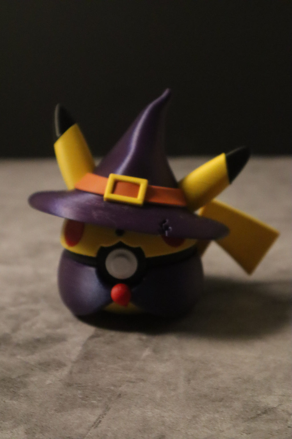 A Pikachu-themed toy figure wearing a purple witch hat with a yellow buckle and orange band, standing on a gray surface against a dark background.